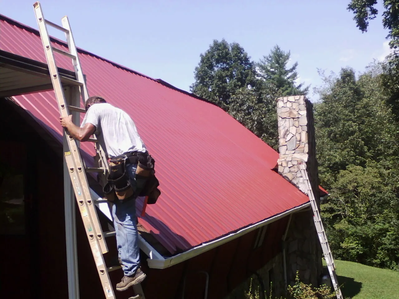 Roofer on a ladder beside a red metal roof and stone chimney — Soffit & Fascia Repair by Roofing Brothers in Kendall